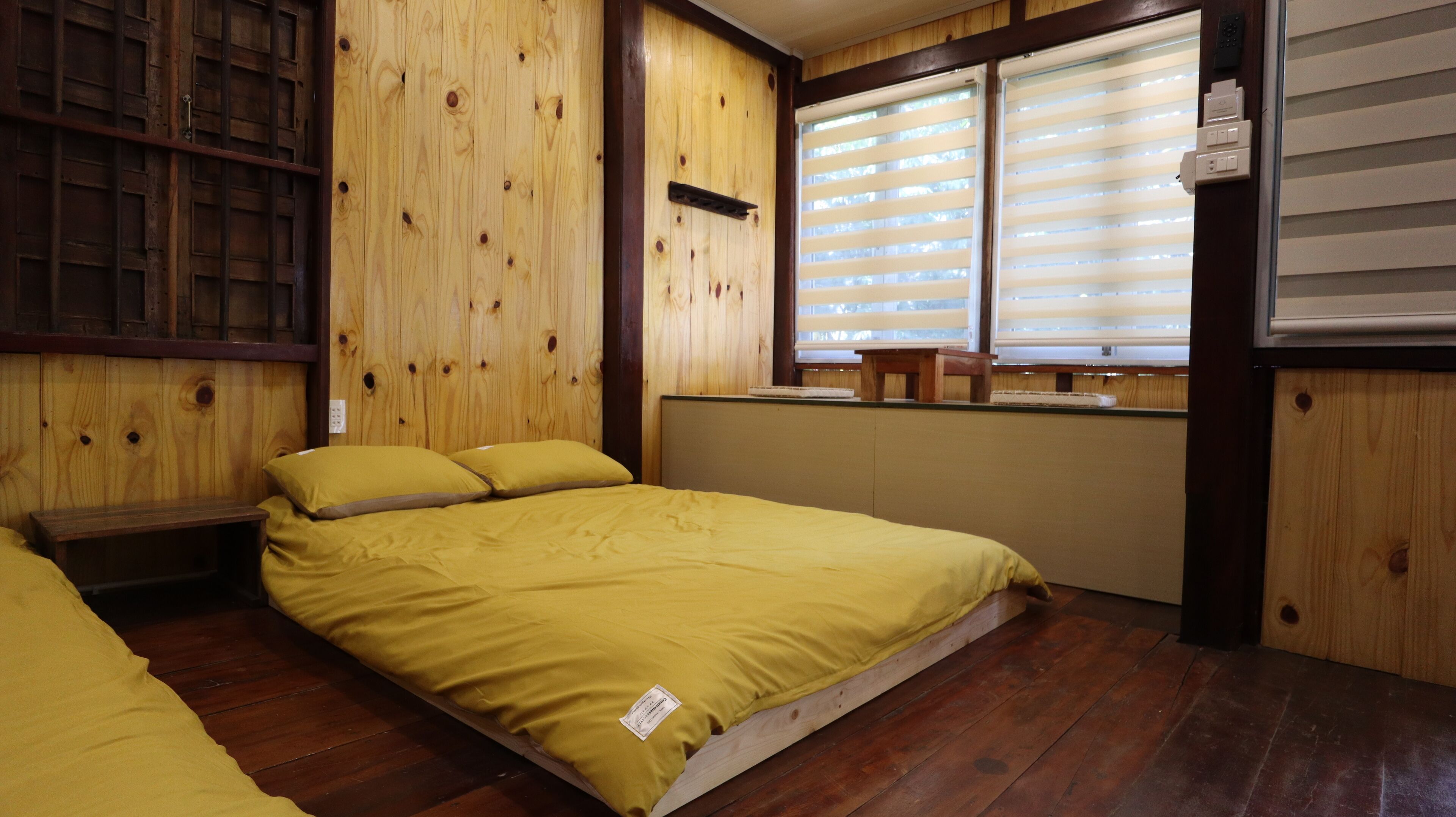 A room with a wooden floor, a bed with yellow bedding, a wooden headboard, a window with blinds, and a small table with a chair.