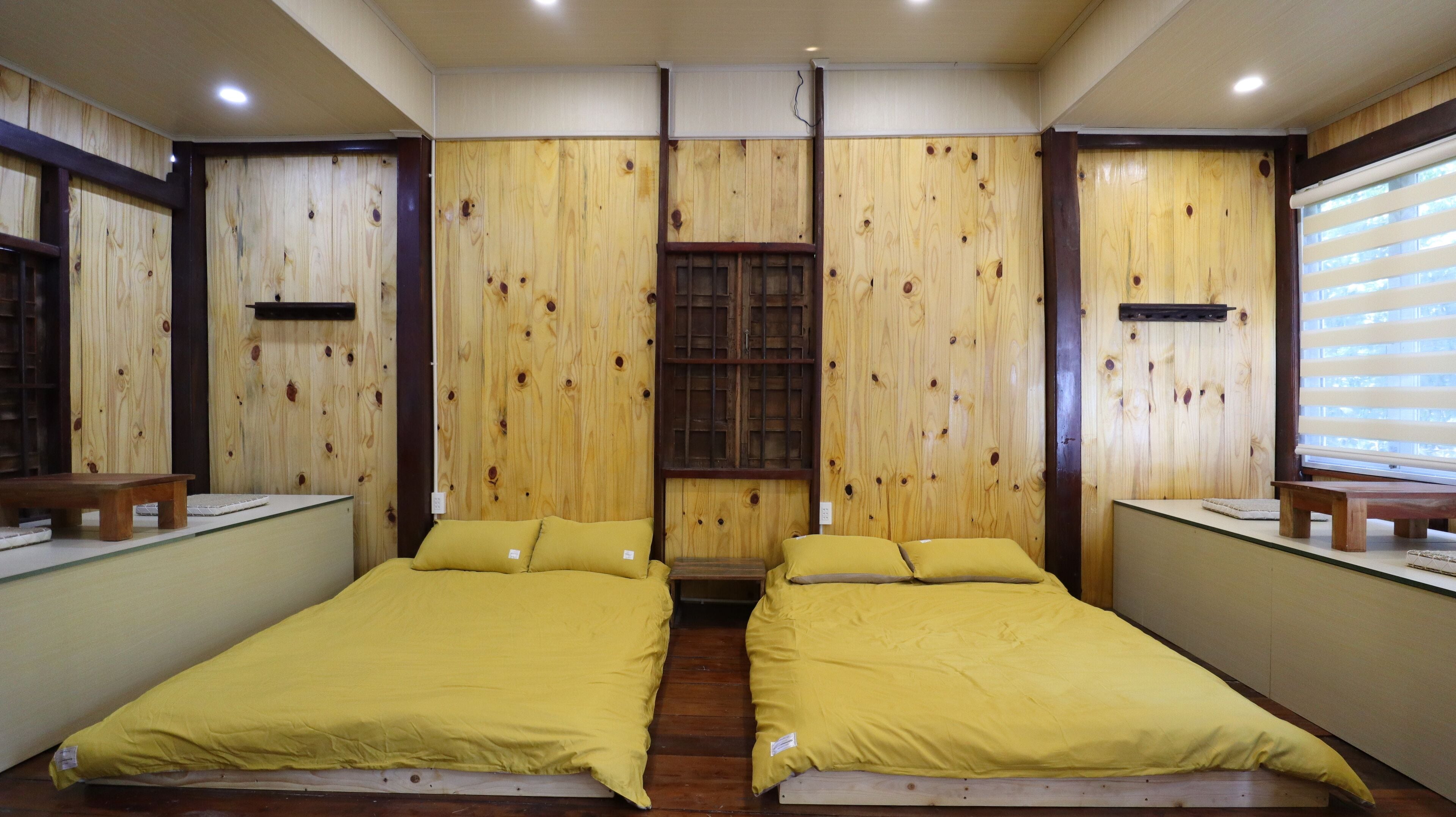 A room with two beds, wooden panel walls, a window with blinds, and a small table.