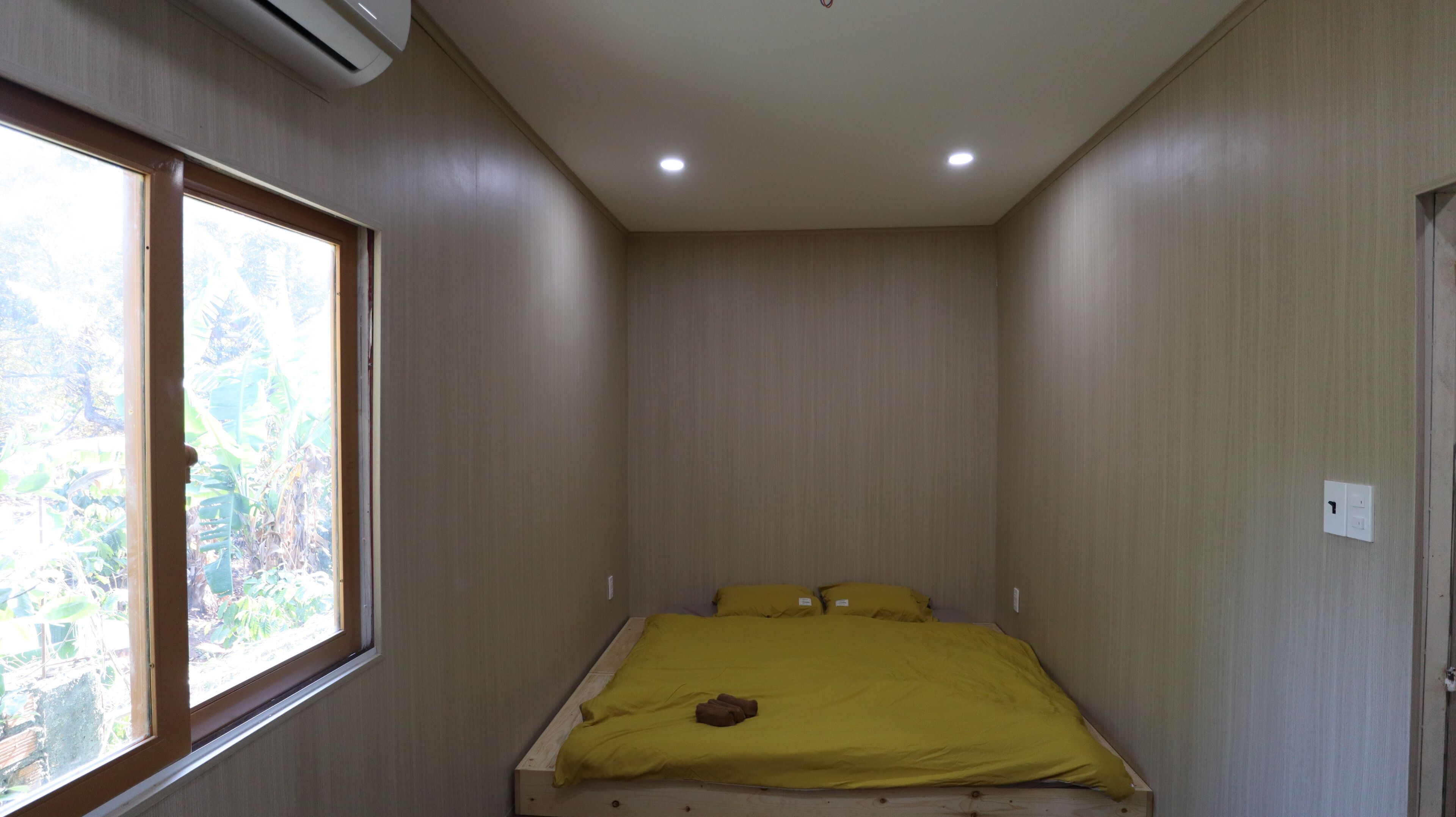 A small, narrow room with a single bed, a window, and an air conditioning unit.