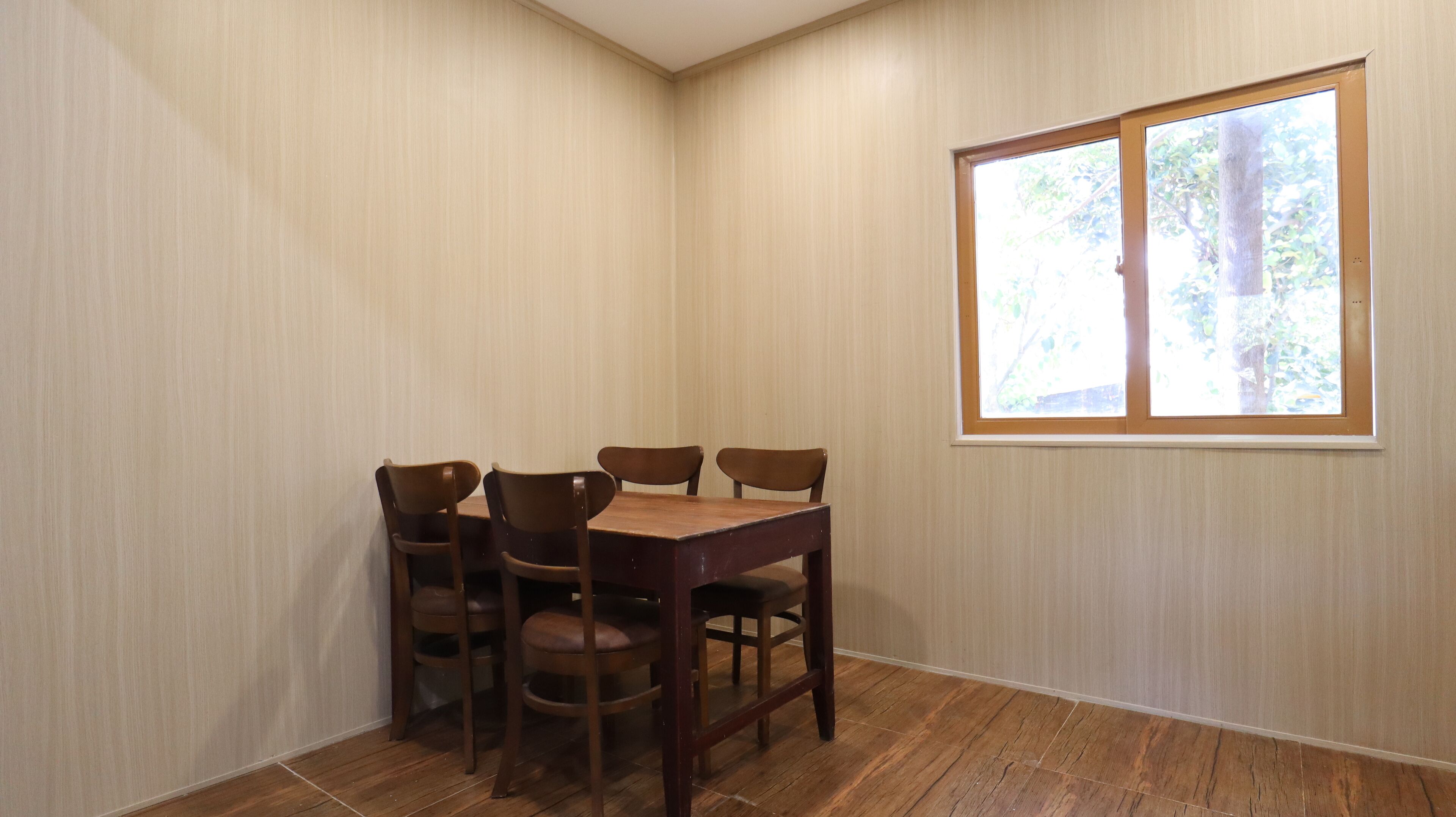 Habitación de esquina con una mesa de comedor y cuatro sillas, una ventana con vistas a árboles, y suelo de madera.