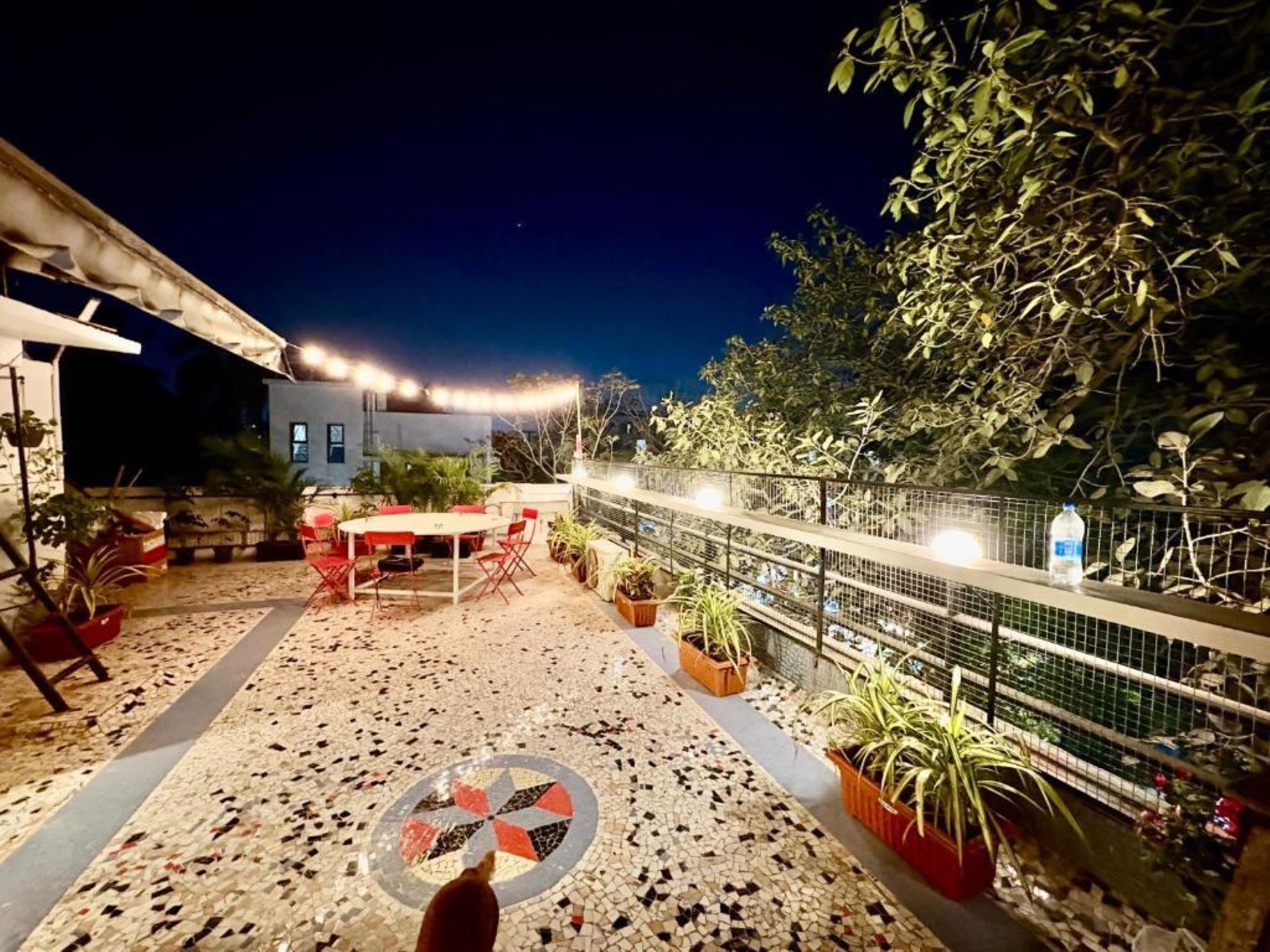 Un patio al aire libre con suelo de baldosas, una mesa redonda y sillas. Hay plantas en macetas y una valla de metal.