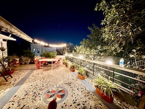 An outdoor patio area with a tiled floor, a round table, and chairs. There are potted plants and a metal fence. - Locomo Hostel Mini - 9 mins from airport (Mumbai)
