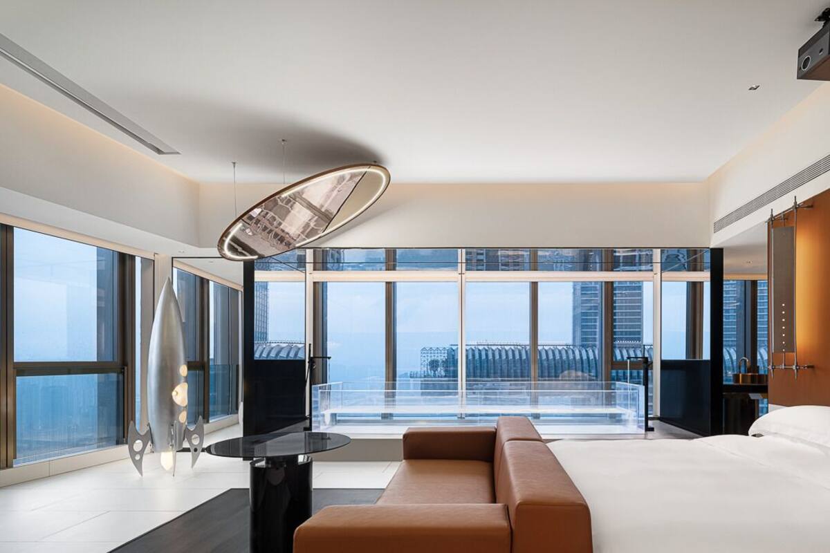 Ein modernes Hotelzimmer mit einem großen Bett, einer Sitzecke und Blick auf die Stadt durch raumhohe Fenster.