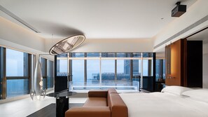 Quarto de hotel moderno com uma cama grande, área de estar e vista da cidade através de janelas do chão ao teto.