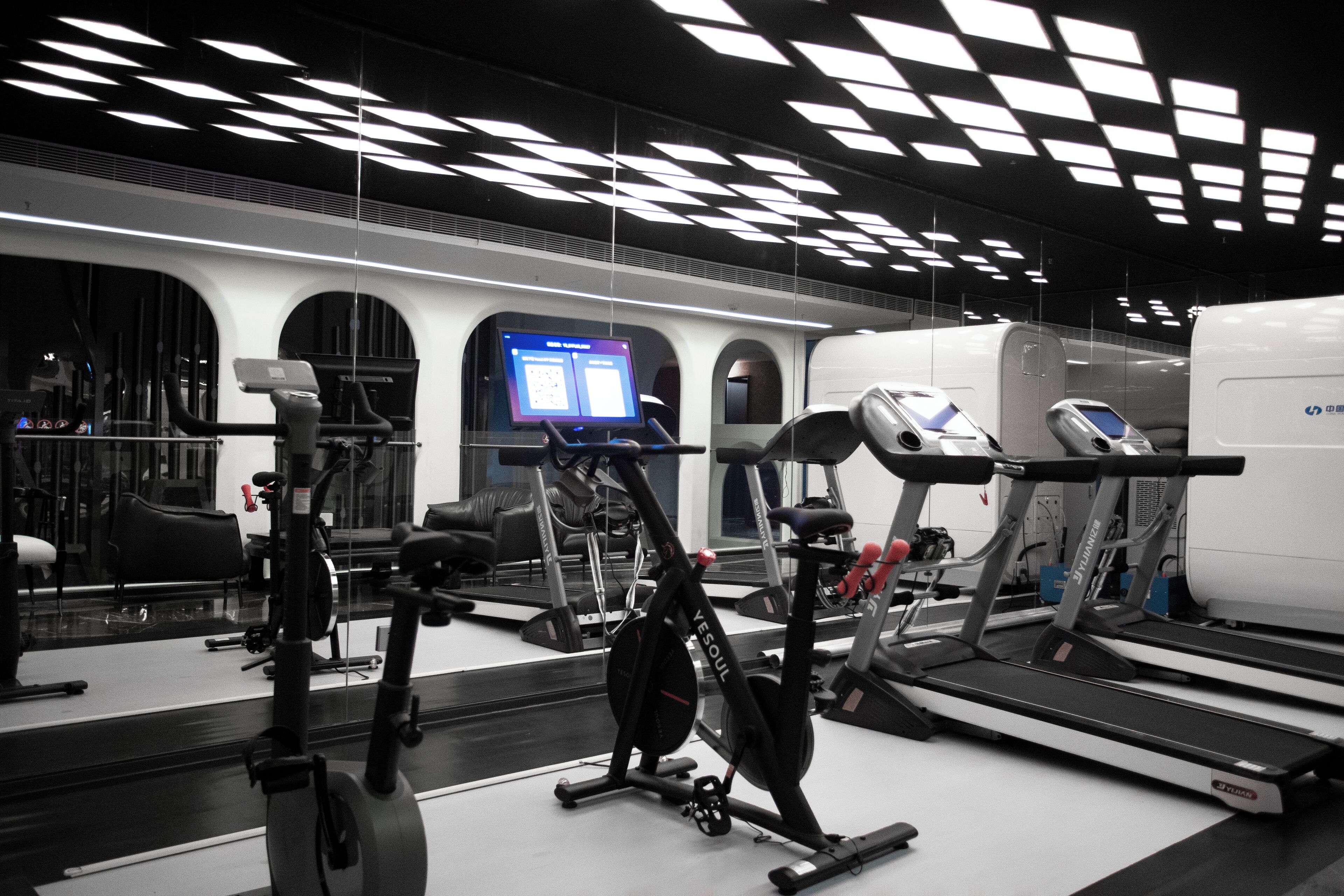 A modern gym with various exercise equipment, including treadmills and stationary bikes, and a digital display screen.