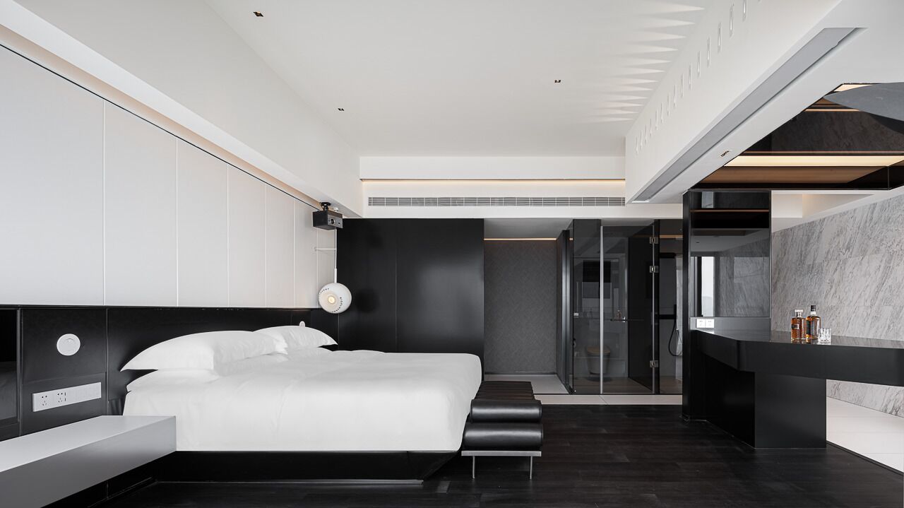 A modern hotel room with a large bed, a bedside table, a built-in desk, and a bathroom with a glass shower.