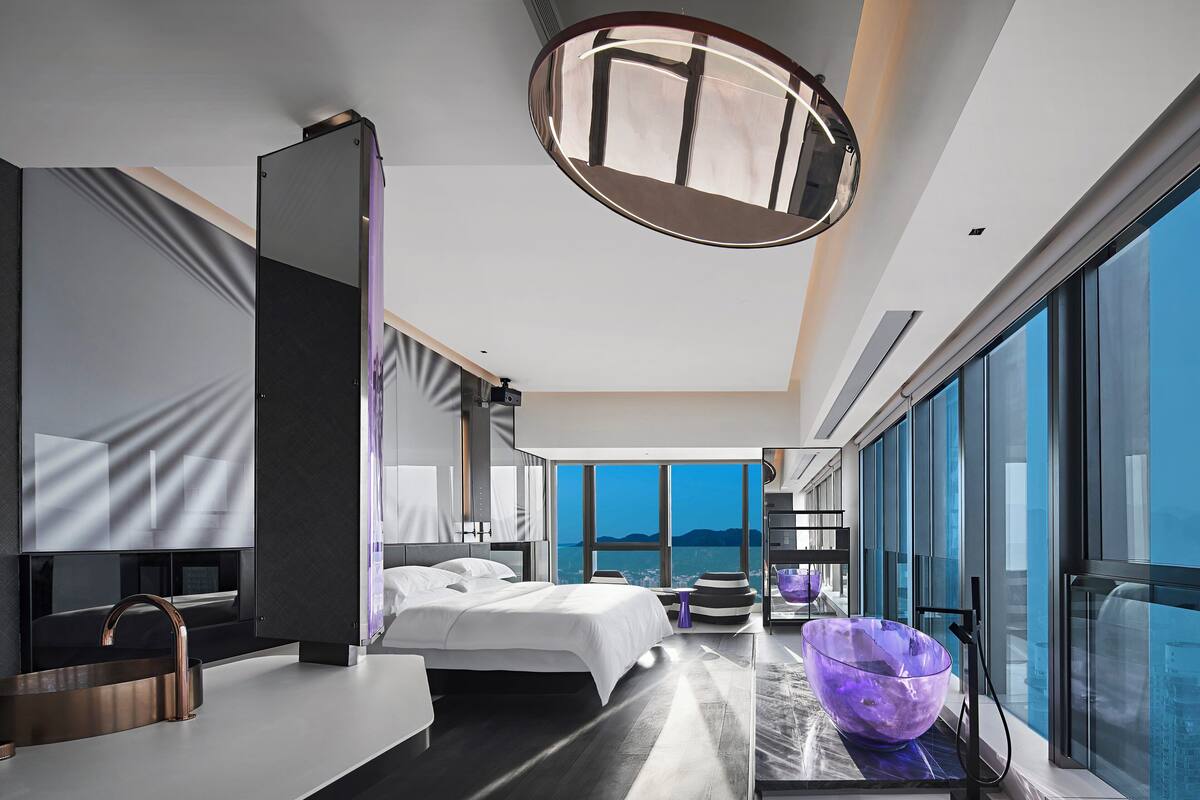Ein modernes Hotelzimmer mit einem großen Bett, einem Badezimmer mit einem modernen Waschbecken und großen Fenstern mit Blick auf die Stadt.