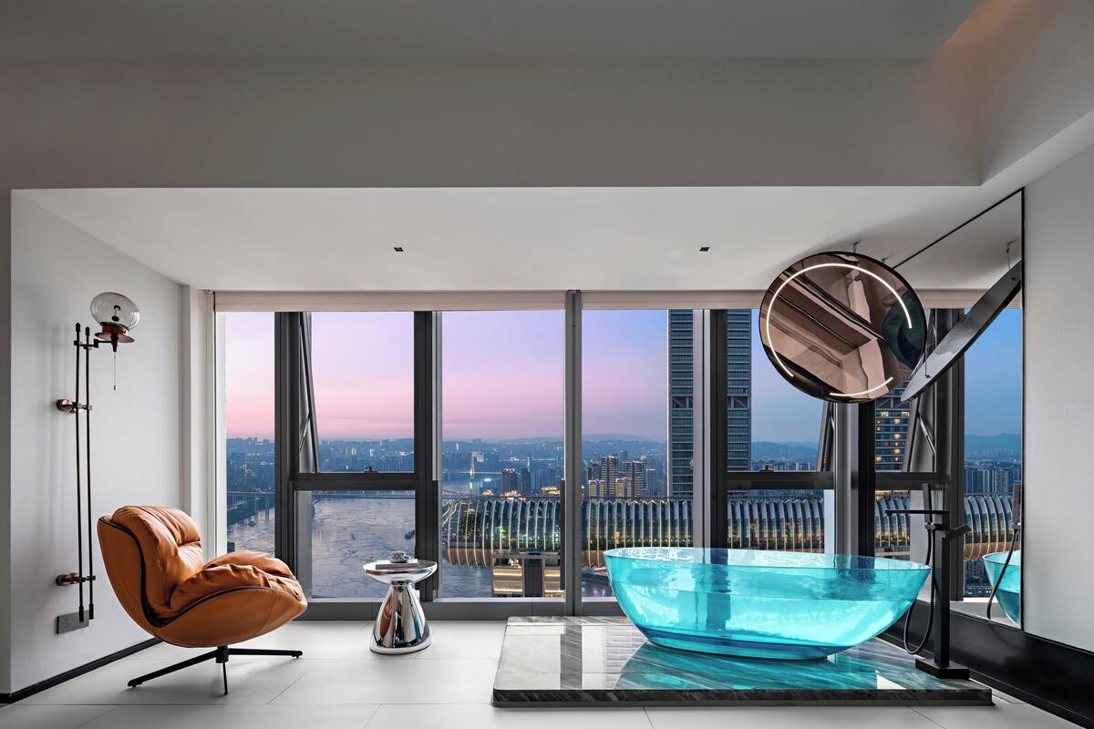 Ein modernes Badezimmer mit großem Fenster, einer Glasbadewanne, einem Ledersessel und einem kleinen Tisch.