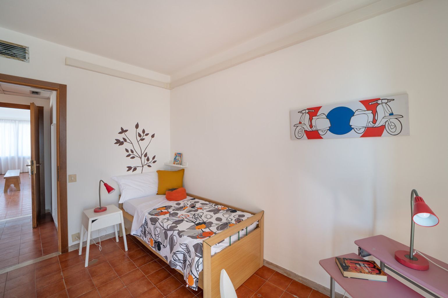Ein kleines, ordentliches Schlafzimmer mit einem Einzelbett, einem Nachttisch, einem Stuhl und einem Schreibtisch. Ein an der Wand hängendes Kunstwerk zeigt einen Roller, und eine Blätter-Abziehbild ist ebenfalls vorhanden.