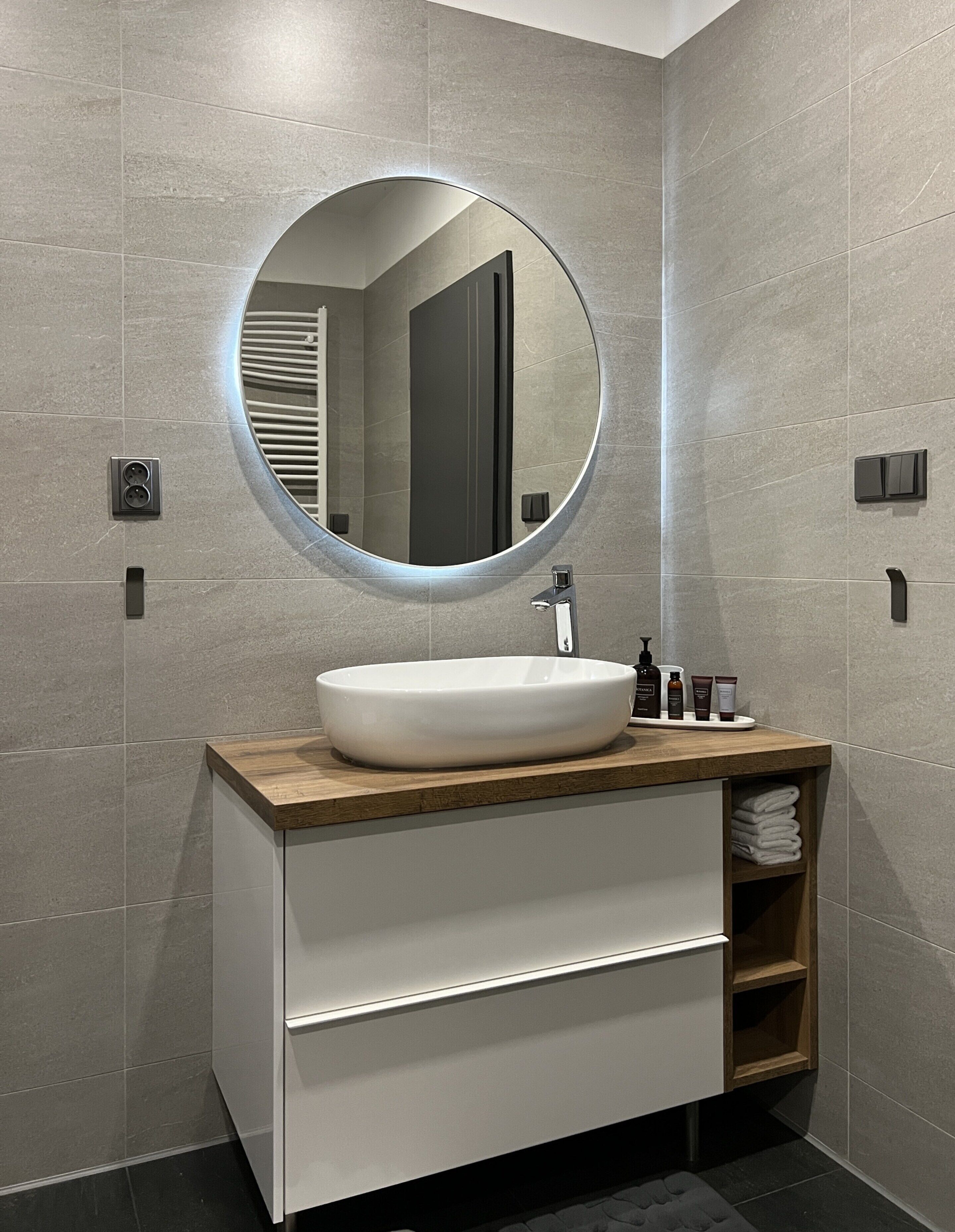 A modern bathroom with a round mirror, a white sink, and a wooden vanity.
