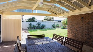 An outdoor patio with a wooden table and benches, a covered ceiling, and a view of a backyard with a brick wall and plants.