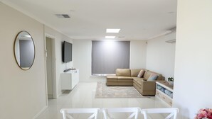 A modern living room with a beige sofa, a flat-screen TV, a dining table with chairs, and a large mirror on the wall.