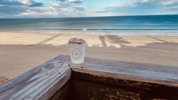 A wooden bench with a coffee cup on it, overlooking a beach with the ocean and a cloudy sky.