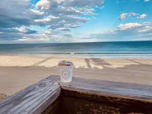 A wooden bench with a coffee cup on it, overlooking a beach with the ocean and a cloudy sky.