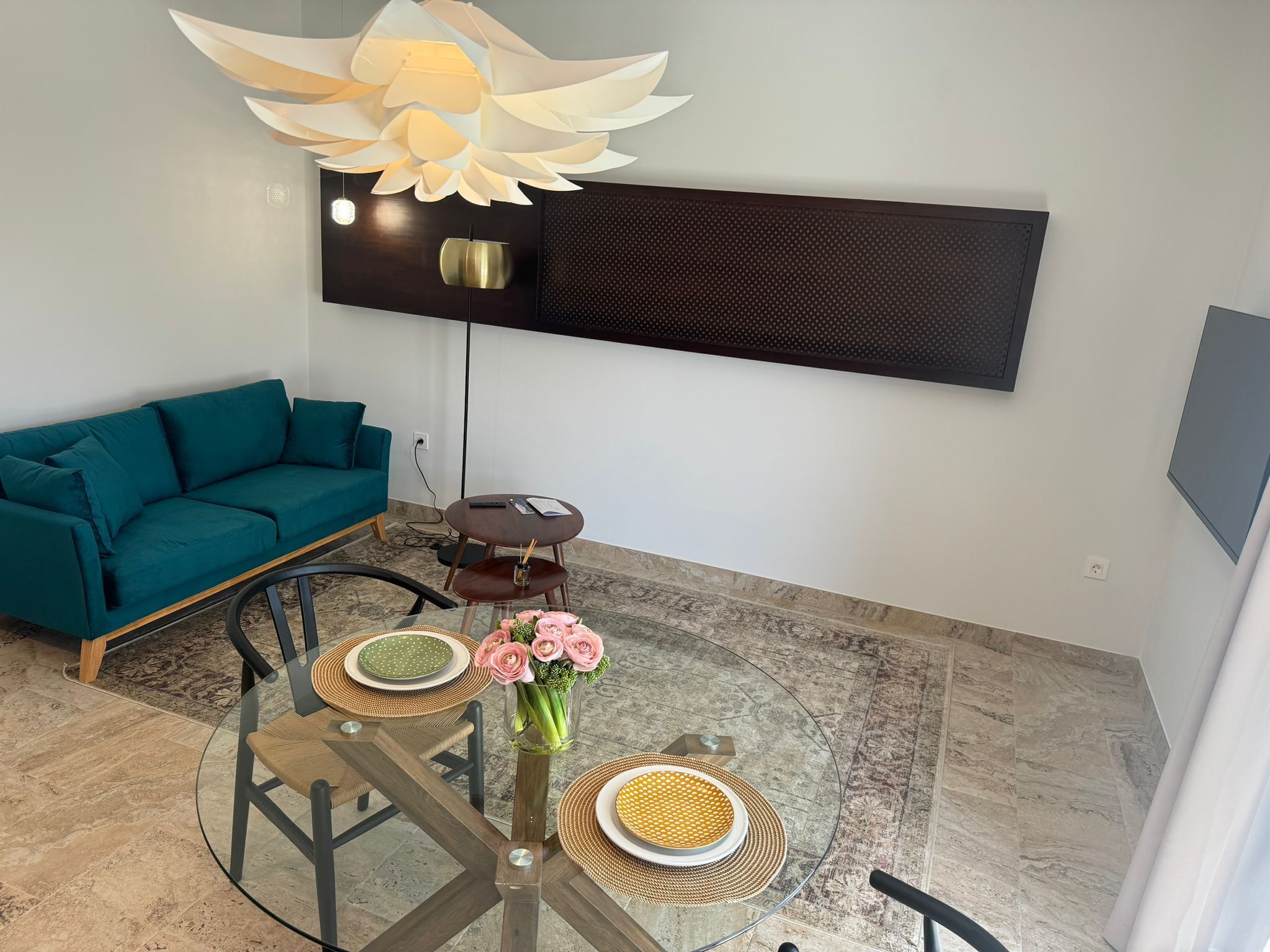 Ein modernes Wohnzimmer mit einem petrolfarbenen Sofa, einem Glastisch mit Untersetzern, einer Blumenvase und einem großen, an der Wand montierten Fernseher.