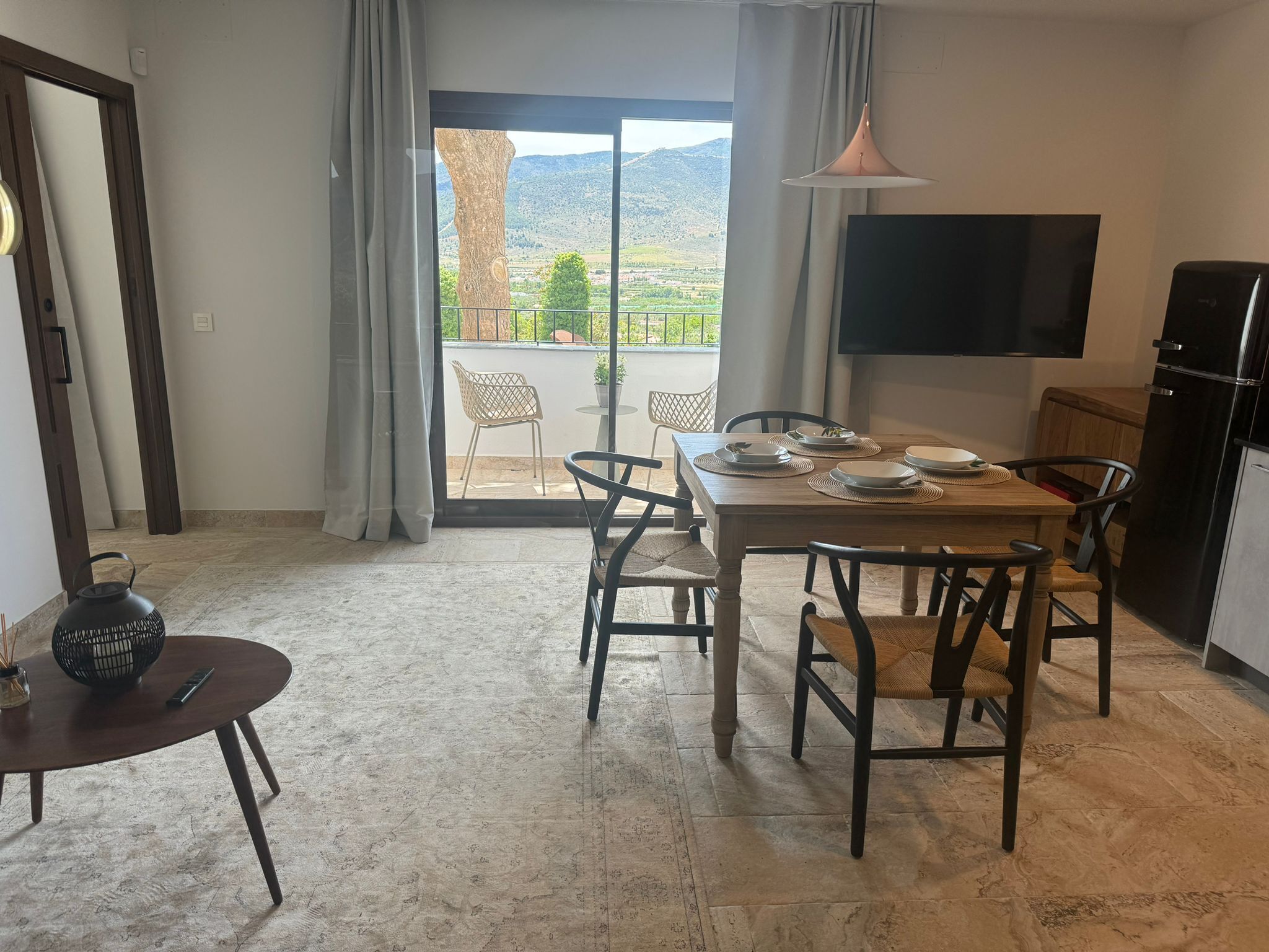 A dining area with a table set for four, a television mounted on the wall, and a view of a mountainous landscape through a glass door.