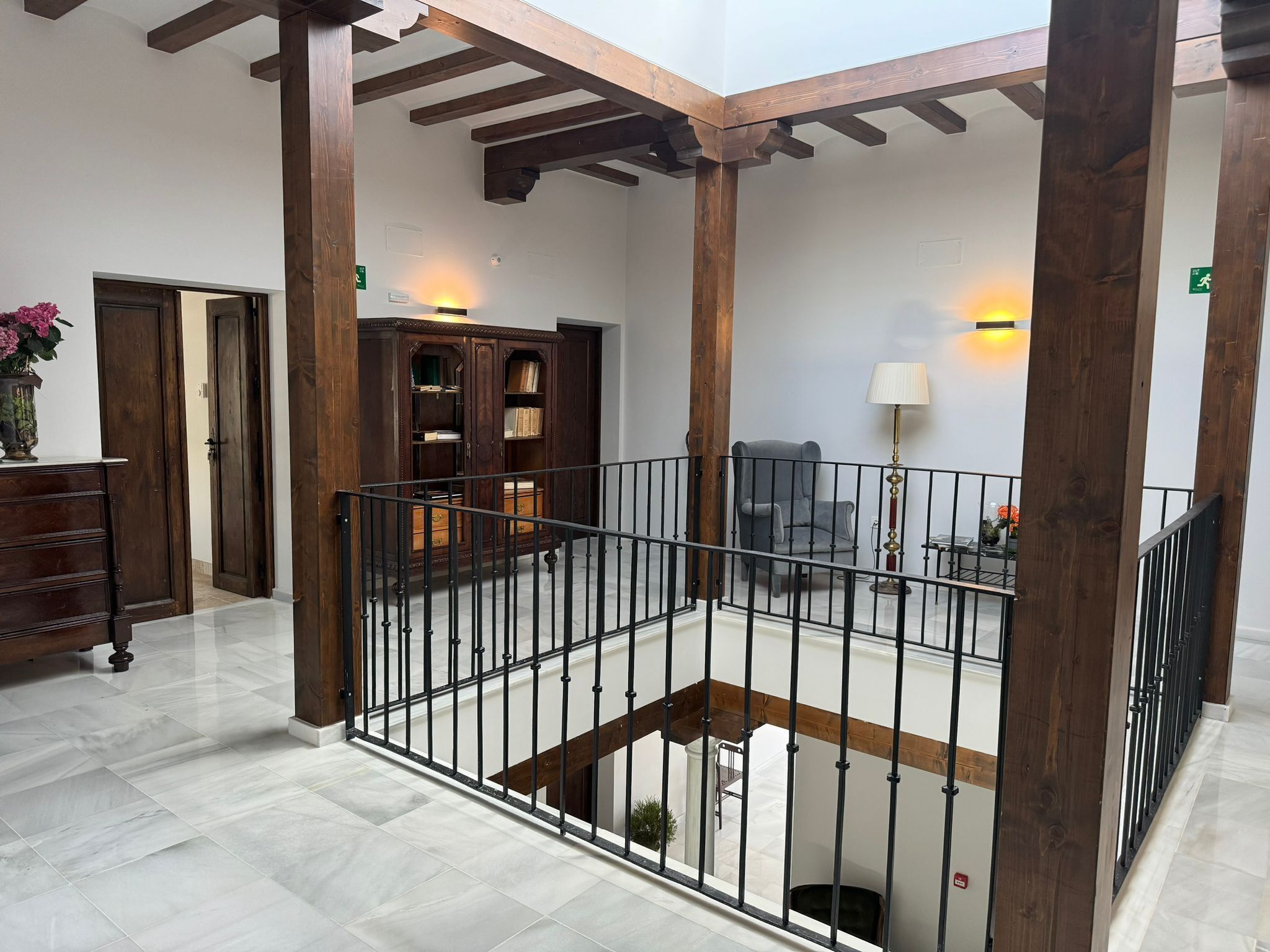 A spacious indoor area with a high ceiling, wooden beams, a metal railing, a bookshelf, a chair, and a lamp.