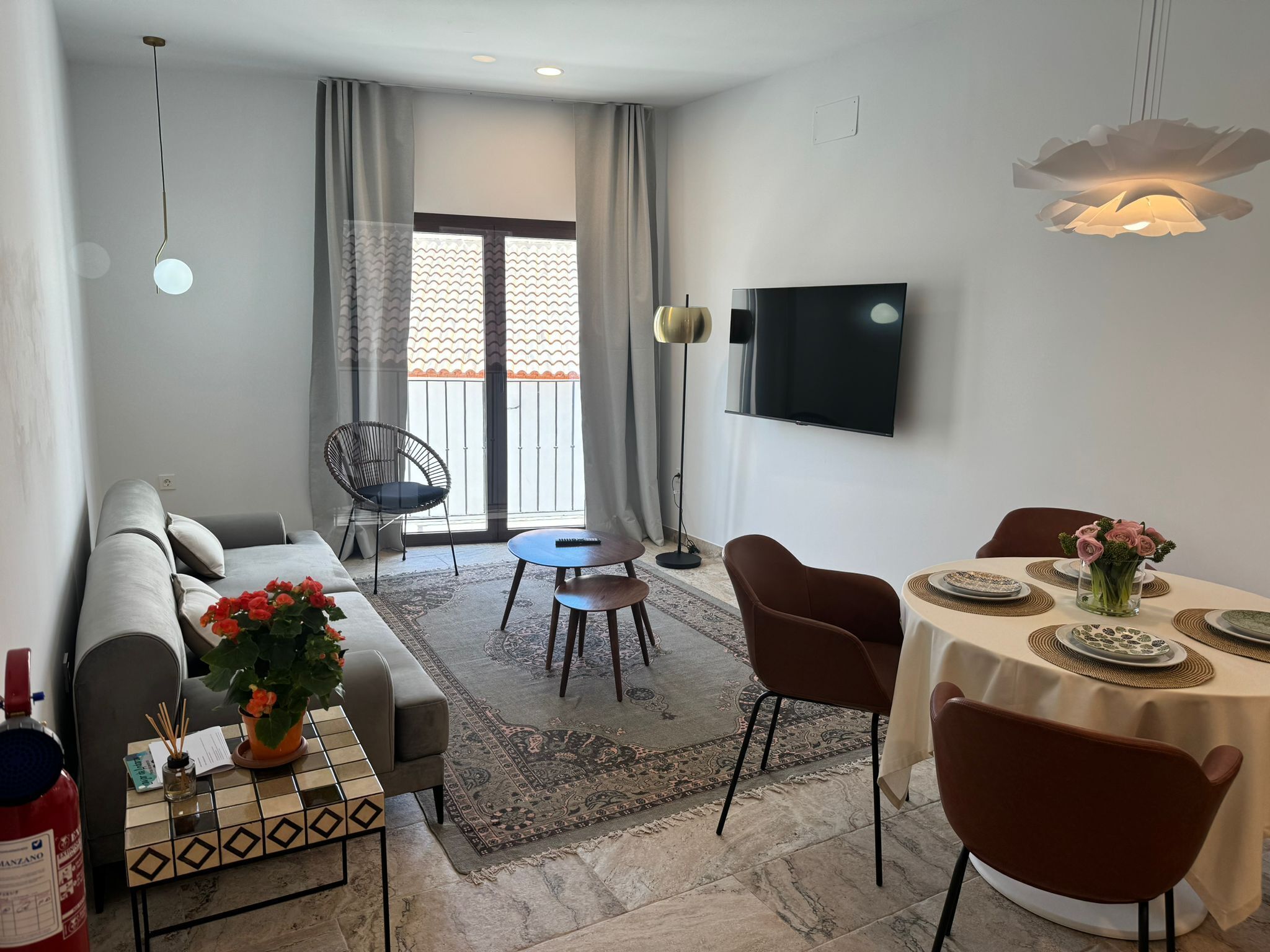 Ein modernes Wohnzimmer mit einer Couch, einem Couchtisch, einem Esstisch mit Stühlen, einem an der Wand montierten Fernseher und einem großen Fenster mit Vorhängen.