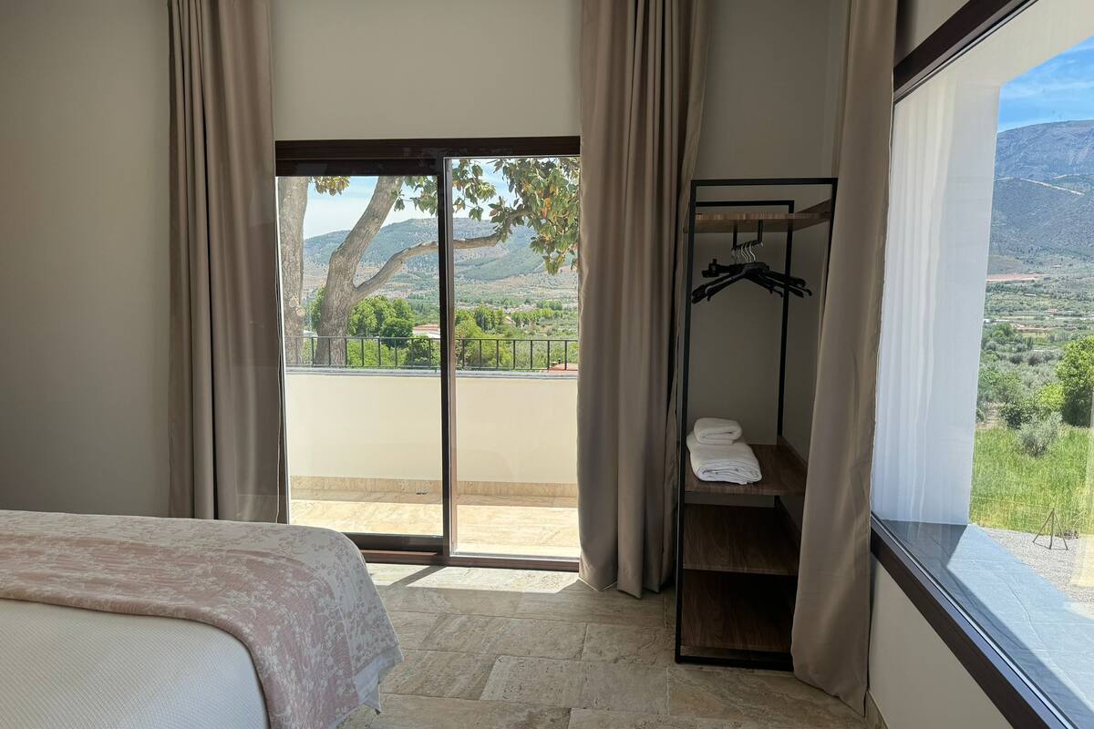 A bedroom with a bed, a wooden shelf with towels, and a view of a mountainous landscape through a large window.