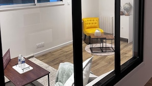 Un intérieur moderne avec une grande fenêtre, un fauteuil jaune, une petite table avec une bouteille d’eau et un radiateur blanc.