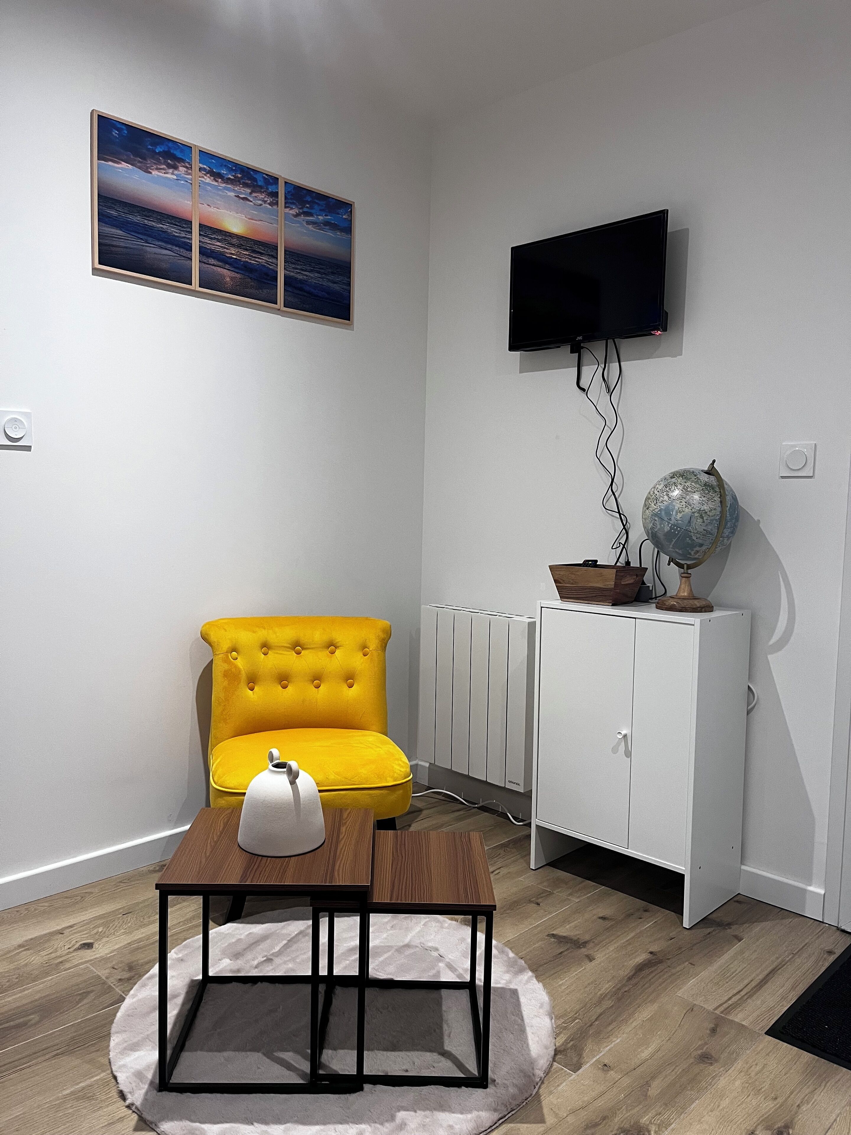 Un soggiorno moderno con una poltrona gialla, un tavolino in legno con una lampada, un termosifone bianco, una TV montata a parete e un mappamondo su un mobiletto.