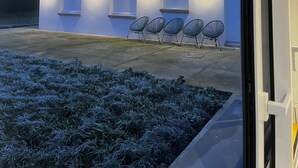 Un bâtiment moderne doté d’un toit plat, de quatre fenêtres avec volets et d’une rangée de quatre chaises noires devant l’entrée.