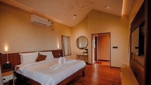 Quarto de hotel com uma cama grande, piso de madeira, ventilador de teto, ar condicionado, mesa de cabeceira, espelho e guarda-roupa.