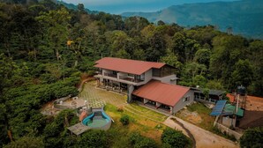 Uma casa de dois andares com telhado vermelho, cercada por vegetação exuberante e uma piscina.