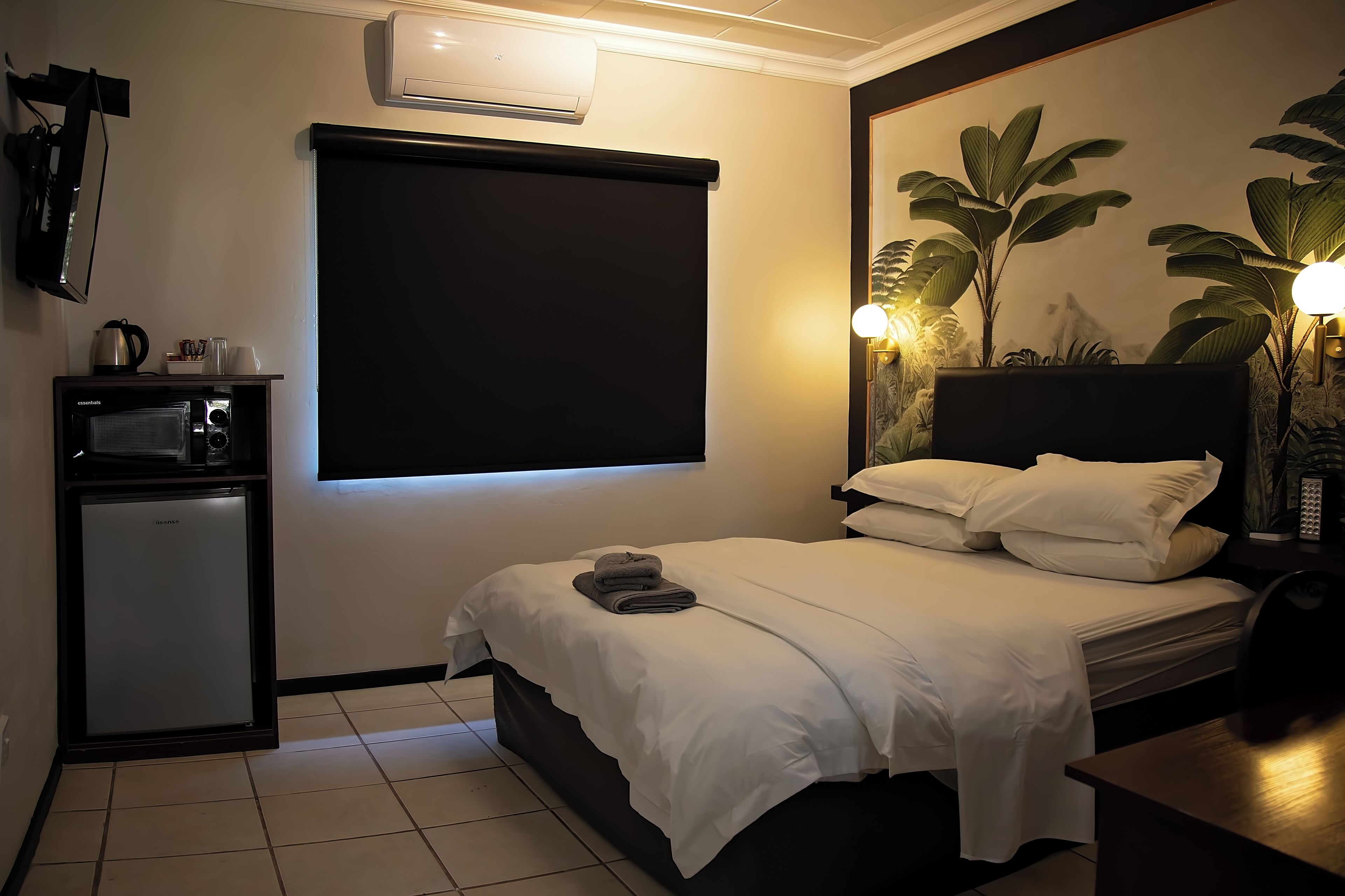 A compact hotel room with a bed, a small kitchenette, and a wall-mounted air conditioner.