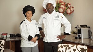 Two chefs in white uniforms standing in a kitchen with a coffee machine and a vase of flowers in the background. - Stay (Tsantsabane)