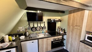 A compact kitchen with a dishwasher, a microwave, and a stove. There are hanging pots and pans, a sink, and a flat-screen TV mounted above the counter.
