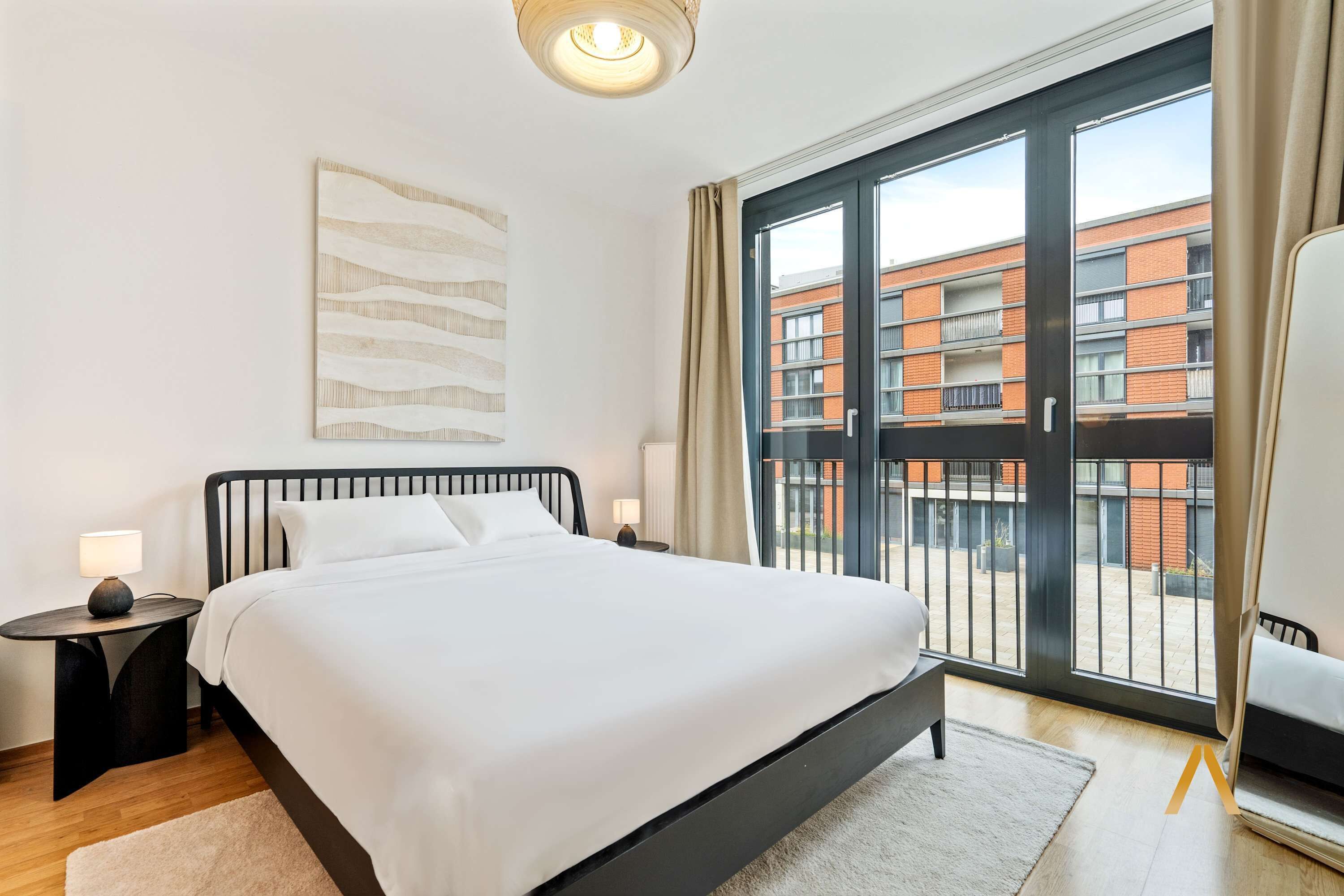 A modern hotel room with a large bed, a nightstand, a lamp, and a view of a building through a glass door.
