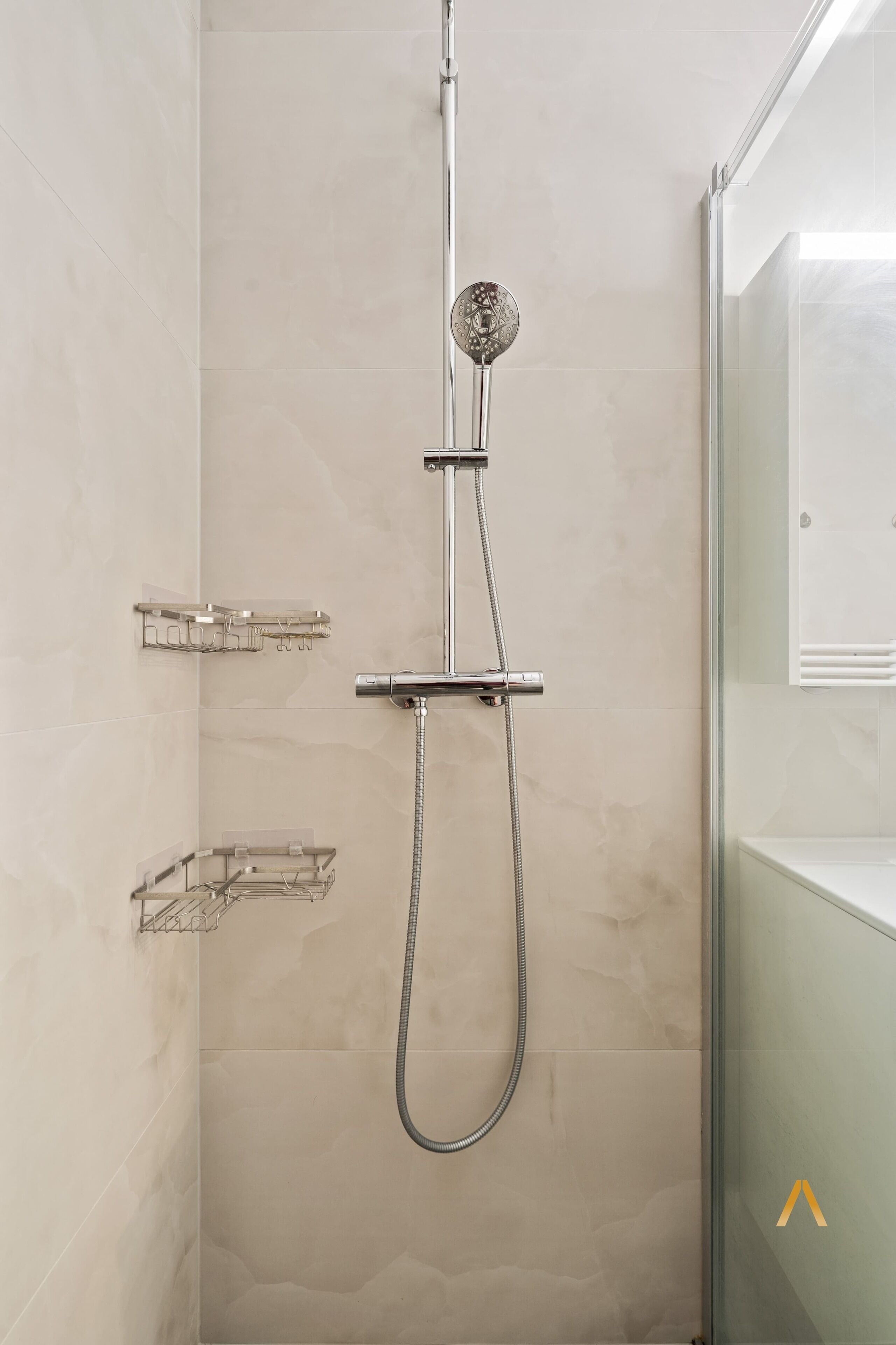 A shower with a handheld showerhead, a wall-mounted faucet, and two clear glass shelves.