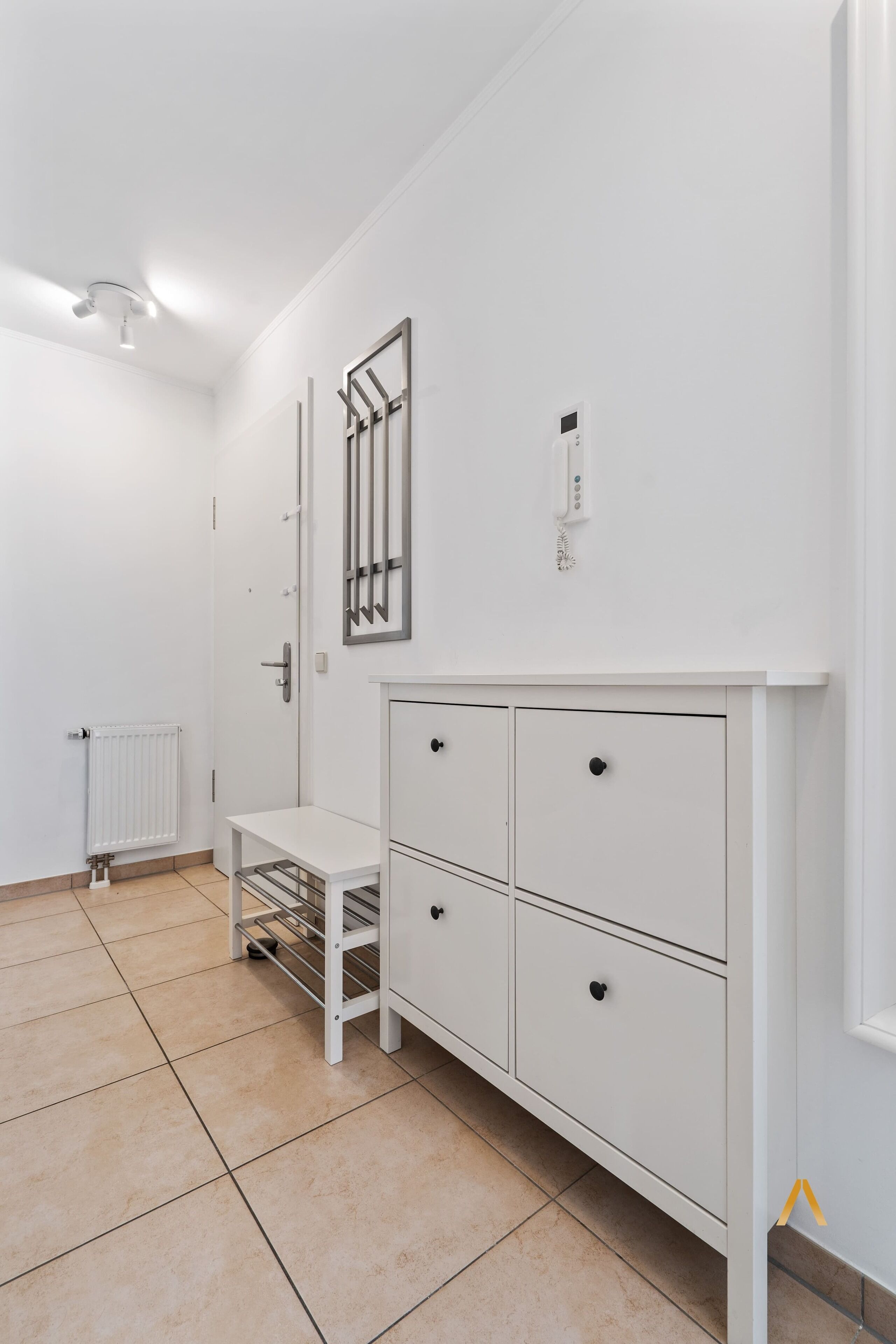 Ein weißer Raum mit Heizkörper, einem weißen Schrank mit Schubladen, einem kleinen Tisch mit einem Metallgestell und einer an der Wand befestigten Leuchte.