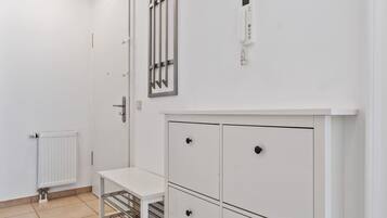Ein weißer Raum mit Heizkörper, einem weißen Schrank mit Schubladen, einem kleinen Tisch mit einem Metallgestell und einer an der Wand befestigten Leuchte.