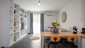 Una cocina moderna con una barra de madera, taburetes negros y una mesa de comedor. A la izquierda, hay una estantería con varios objetos, y una ventana grande con cortinas.
