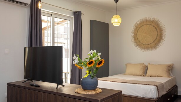 Un dormitorio con televisión sobre un mueble de madera, una cama con ropa de cama color beige, un florero con flores y un adorno circular fijado a la pared.