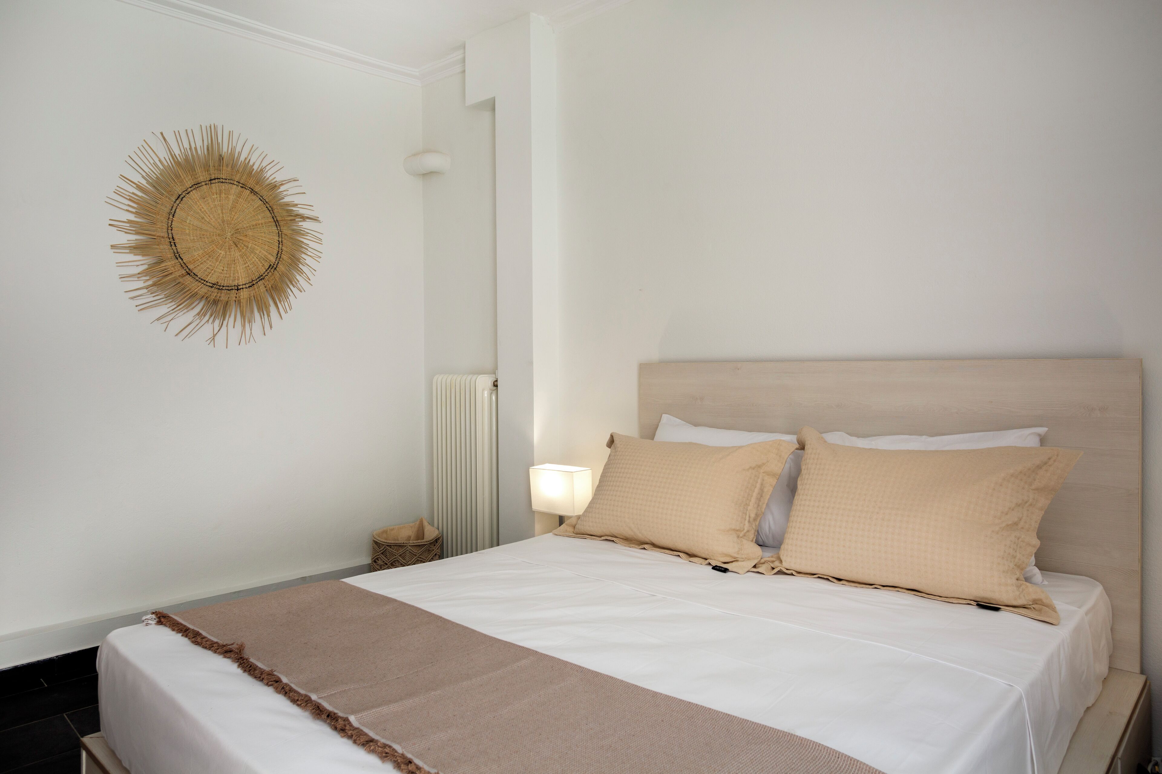 Ein ordentlich bezogenes Bett mit beige farbenem Kopfteil und Kissen, einer Nachttischlampe sowie einer Wanddekoration im Sonnenstrahl-Design.