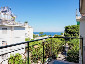 A balcony with a view of the sea, a building with a white facade, and a tree-lined street.