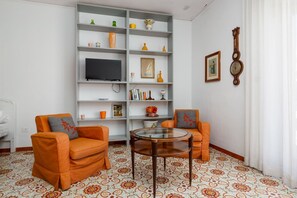Sala de estar com dois poltronas laranja, uma mesa de centro de vidro, uma televisão em uma prateleira branca e ladrilhos de chão estampados.