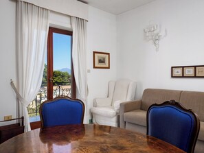 A room with a dining table, two chairs, a sofa, a window with curtains, a framed picture, and a wall-mounted light fixture.