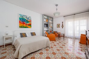 A hotel room with a bed, a desk, a chair, a bookshelf, and a painting on the wall.