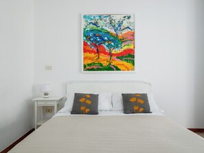 Un dormitorio con una cama, dos almohadas con diseños florales, una mesita de noche con una lámpara y un cuadro abstracto y colorido en la pared.