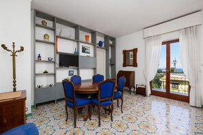 Uma área de refeições com uma mesa redonda e cadeiras azuis, uma TV na parede e uma varanda com vista.
