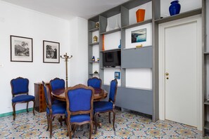 A dining area with a round wooden table and blue upholstered chairs, surrounded by white walls and a tiled floor.