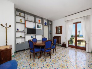 A dining area with a wooden table and blue chairs, a TV on a shelf, and a balcony with a view.