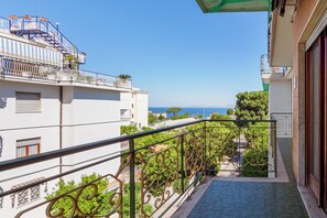 A balcony with a view of the sea, a white building, and a green railing.