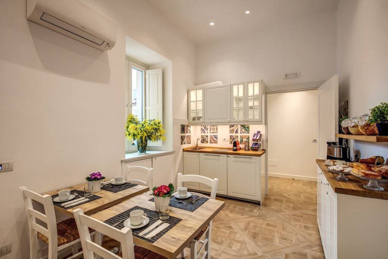 A compact kitchen with white cabinets, a dining table with chairs, and a window with flowers.