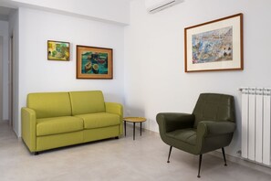 A room with a green sofa, a small round table, and a green armchair. There are three framed artworks on the wall.