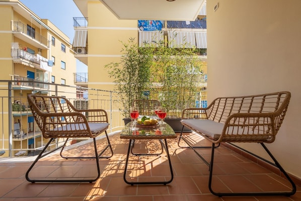 A balcony with two wicker chairs, a glass table with a fruit bowl and glasses, and a potted plant.